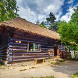 Niebo z chmurą. Zagroda a w niej drewniana chata z podmurówką z kamieni, zbudowana z brązowych beli z niebieskim pasem między nimi, pokryta strzechą, z kominem. Z małymi oknami i wejściem po środku. Po prawej płot ze sztachet, na którym wisi odzież, a za nim widać ogródek z kwiatami i warzywami oraz wysokie drzewo. Po lewej w dali schodki z kamieni wśród zielonej trawy prowadzą do kolejnej chaty.