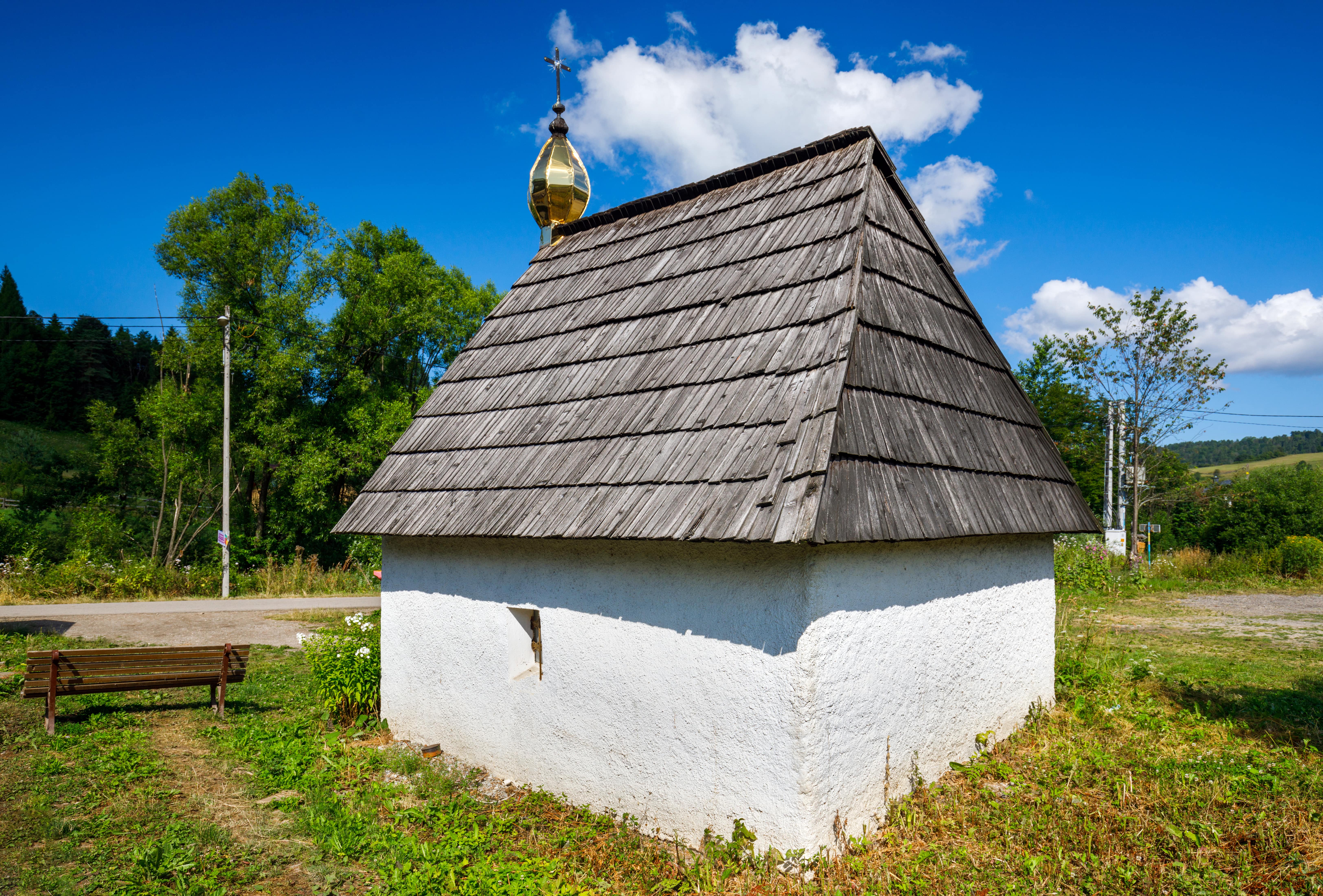 .	Mała, bielona, murowana kapliczka z dachem pokrytym gontem, zakończonym na kalenicy złocistym, baniastym hełmem - widok z tyłu. Przy kapliczce po lewej stoi drewniana ławka. Wokoło trawa. Za kapliczką droga, dalej las. Na niebie kłębiaste chmury.