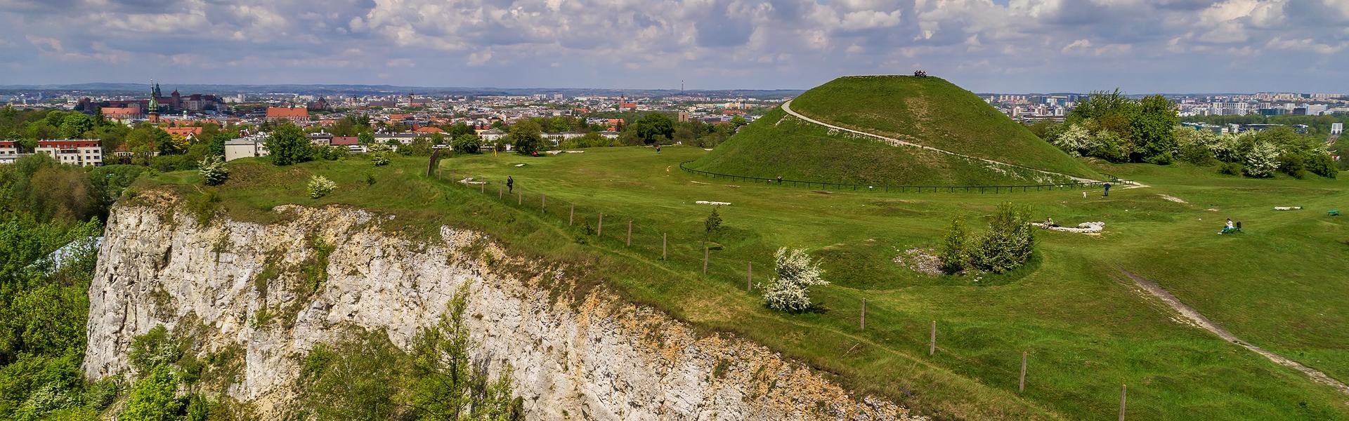 Ein großer, grüner Erdhügel, im Hintergrund ein Blick auf Krakau.