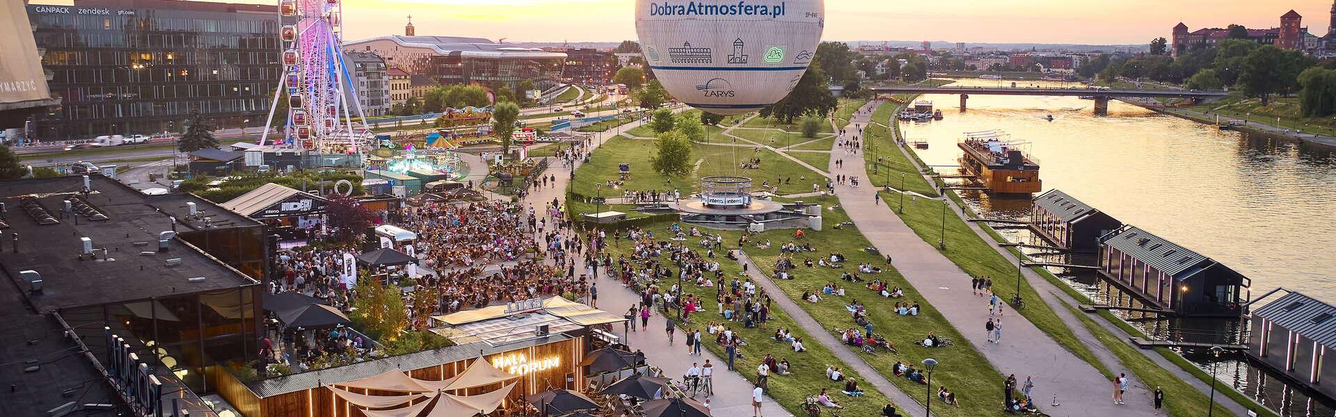 Widok na Forum Przestrzenie w Krakowie z lotu ptaka. Po lewej ustawione parasole obok leżaki i foood tracki. Obok Forum karuzela i balon na ogrzene powietrze. Po prawej rzeka Wisła z zacumowanymi barakmi, w tle Zamek na Wawelu.