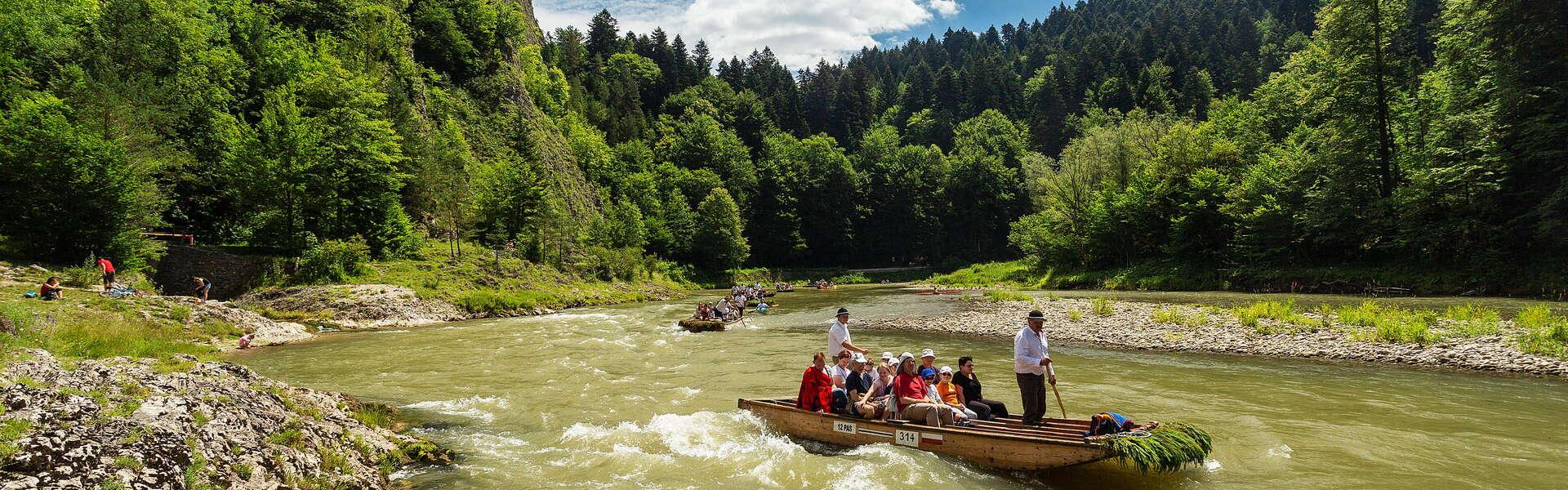 Rafting down the Dunajec River on several rafts. The waves on the Dunajec River are quite high. Limestone rocks form the bank of the river. Three people are sitting on the shore. In the background are the lush Pieniny forest and puffy clouds in the sky.